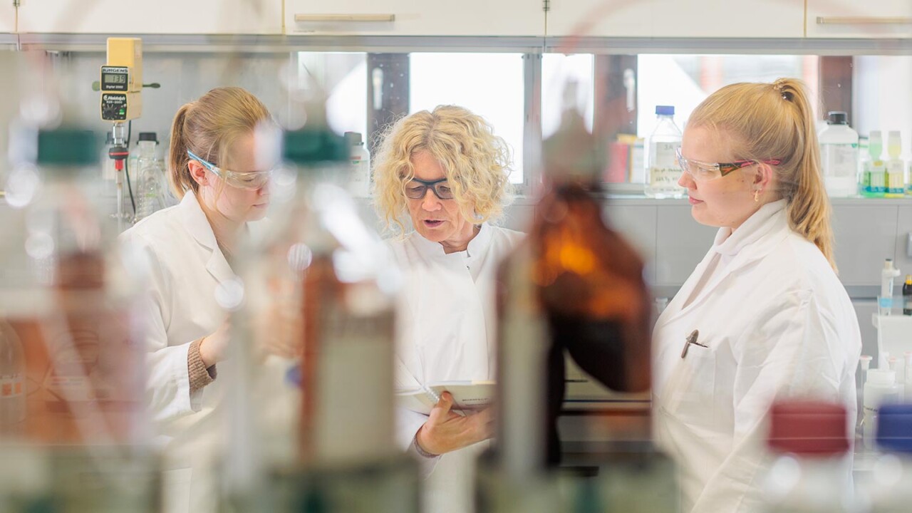 Drei Mitarbeitende in Laborkitteln und Schutzbrillen besprechen Untersuchungsschritte in einem Analytiklabor. Im Vordergrund sind Laborflaschen unscharf zu sehen. Das Bild steht für Zusammenarbeit in der chemisch-analytischen Prüfung zur Lebensmittelauthentizität, Qualitätssicherung und Verfälschungsaufklärung.