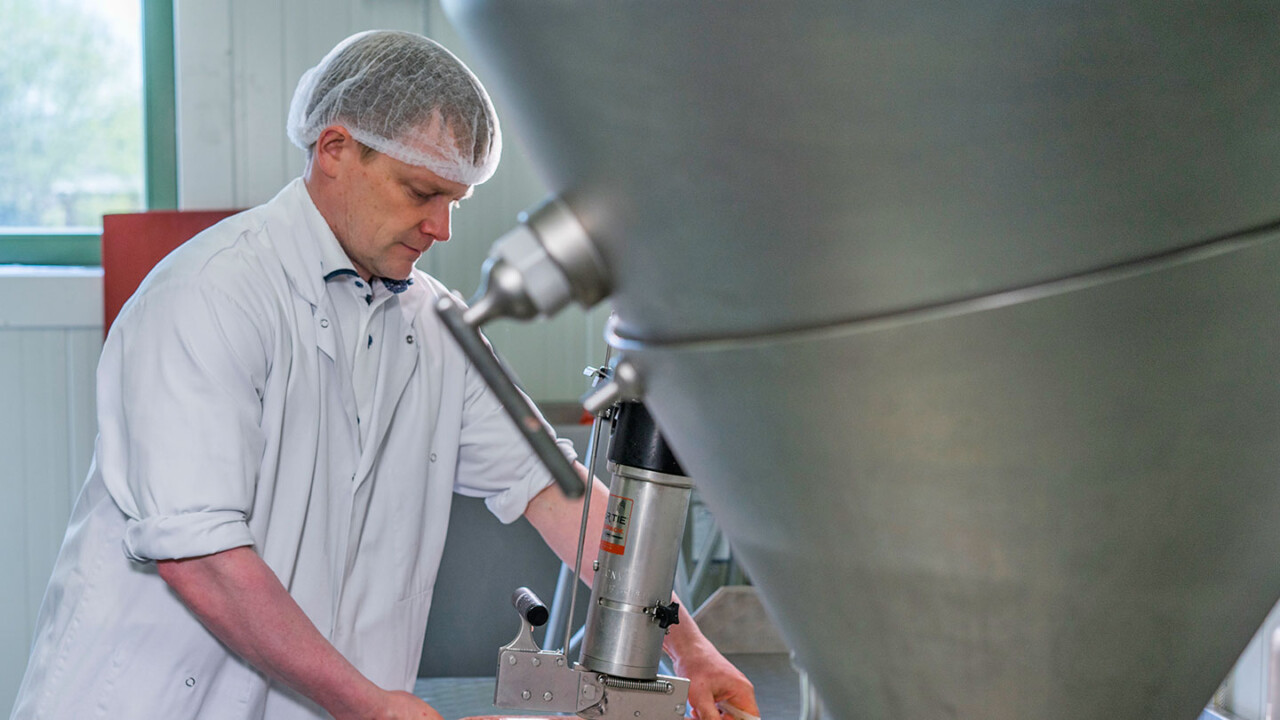 Ein Mitarbeiter in Laborkittel und Haarnetz arbeitet an einer großen Edelstahl-Anlage im Technikum. Er bedient ein Ventil bzw. ein Bauteil an der Anlage und überwacht damit einen biotechnologischen Prozess unter praxisnahen Produktionsbedingungen.