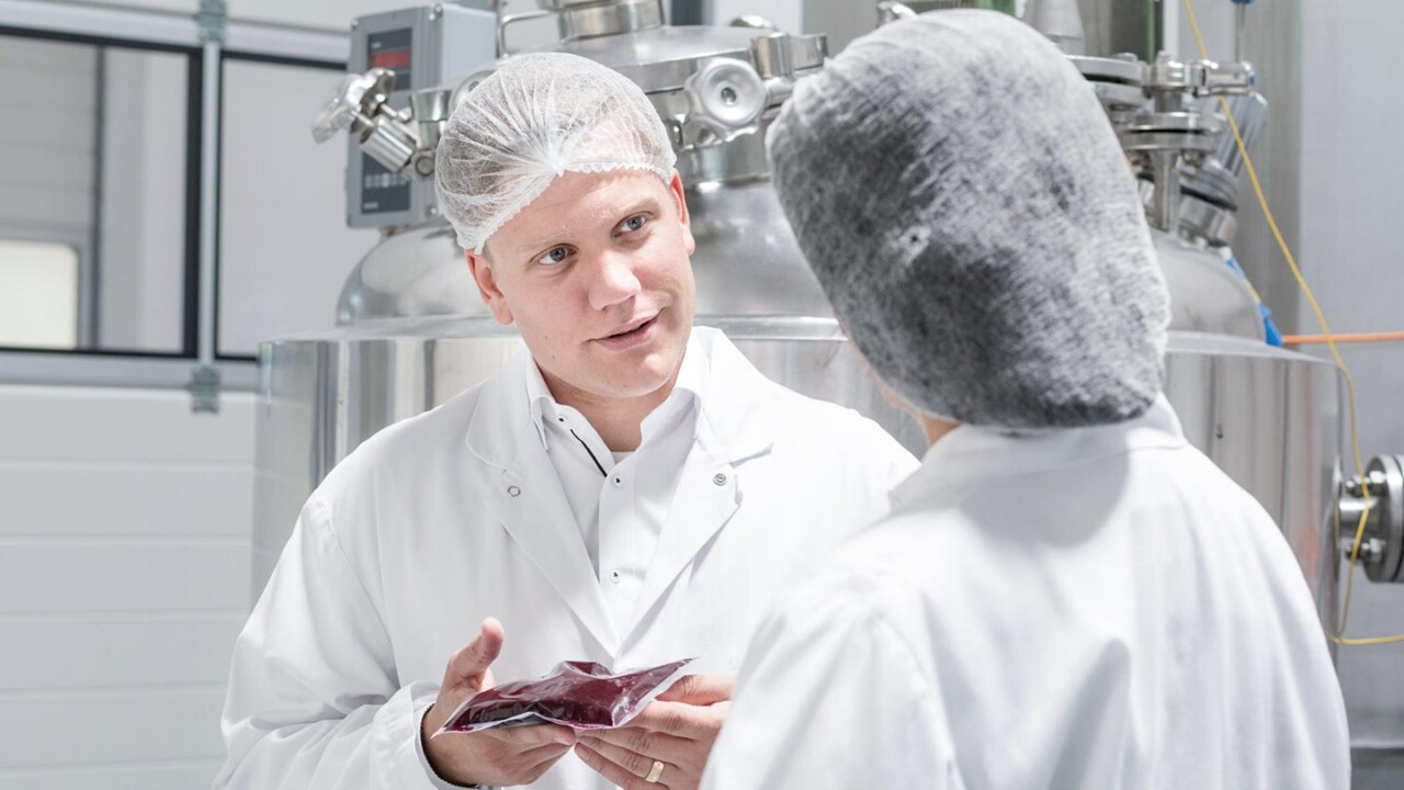 Zwei Mitarbeitende in Laborkitteln und Haarnetzen stehen vor einer Edelstahl-Anlage im Technikum. Eine Person hält eine verpackte Produktprobe in der Hand und bespricht mit der Kollegin bzw. dem Kollegen die nächsten Schritte zur Produktentwicklung und Prozessoptimierung.