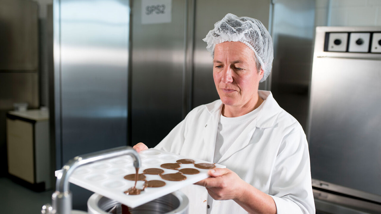 Eine Mitarbeiterin im Laborkittel und Haarnetz arbeitet im Technikum an einer Produktionsanlage und füllt flüssige Schokolade in eine Formplatte. Die Aufnahme zeigt den Prozess der Musterherstellung und Qualitätsprüfung im Rahmen der Produktentwicklung.