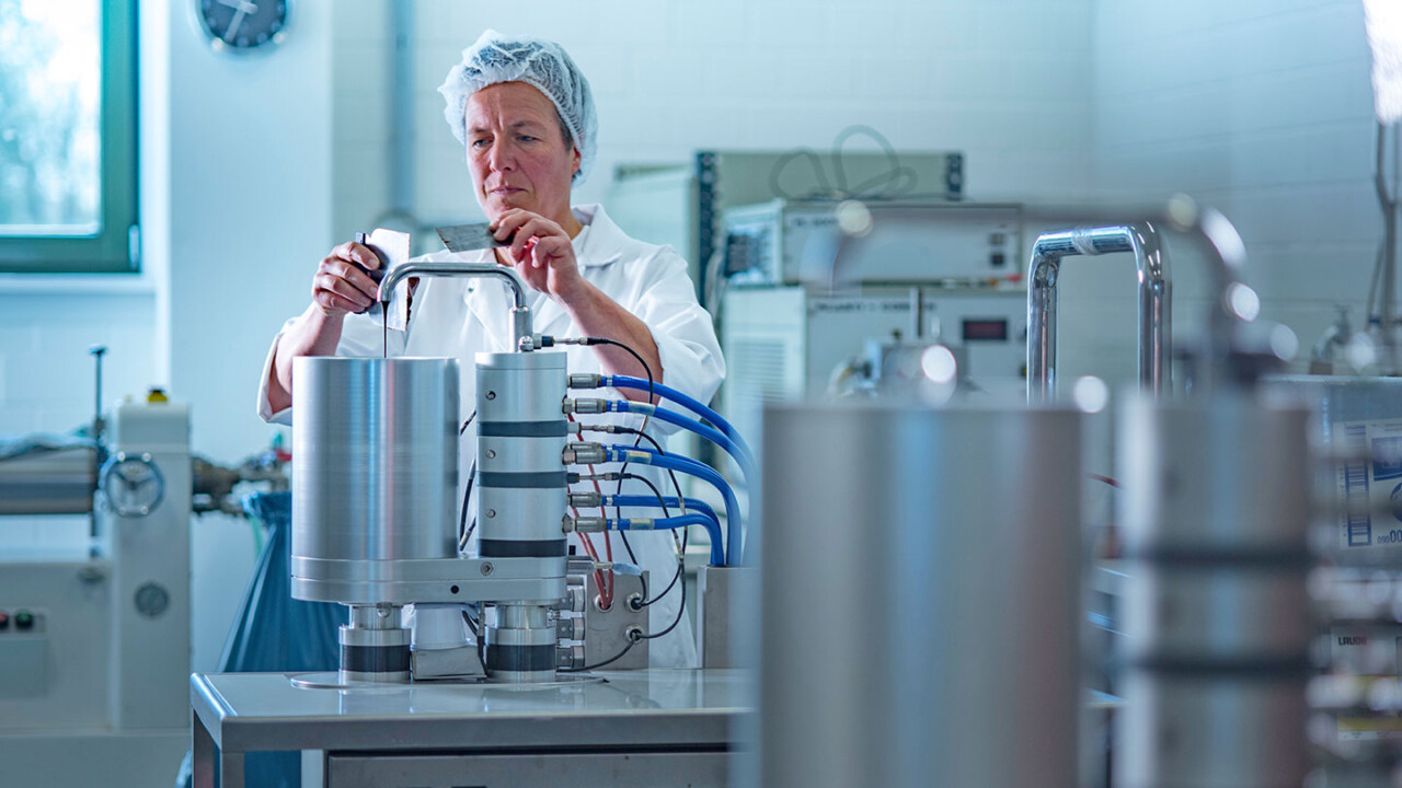 Eine Mitarbeiterin im Laborkittel und Haarnetz bedient eine technische Versuchsanlage im Technikum. Die Aufnahme zeigt die Durchführung und Überwachung eines Prozessschritts im Rahmen der Produkt- und Prozessentwicklung.