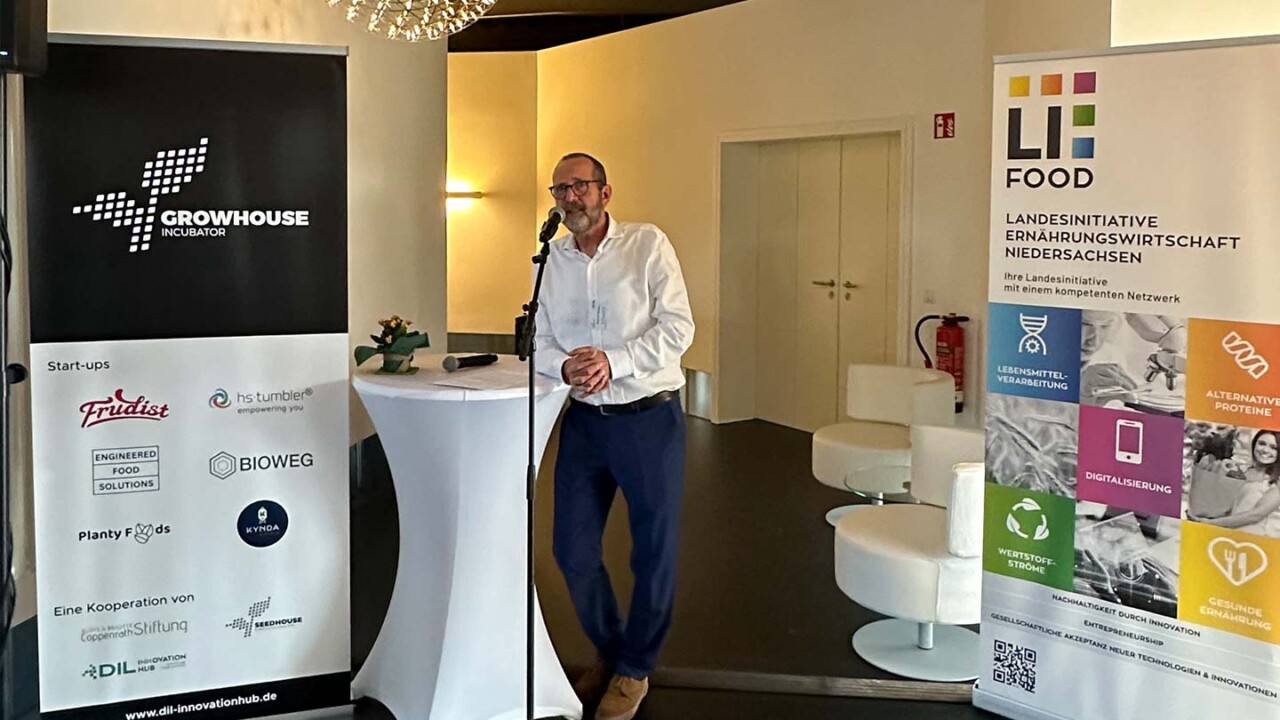 dil-ev-beratung-innovation-hub-Innovationen-und-Start-ups-der-Ernaehrungswirtschaft Ein Referent spricht an einem Stehtisch in einen Mikrofonständer bei einer Veranstaltung im Growhouse Incubator. Neben ihm stehen Roll-up-Banner von „Growhouse Incubator“ und „LiFOOD – Landesinitiative Ernährungswirtschaft Niedersachsen“ mit Themen wie Lebensmittelverarbeitung, Digitalisierung, alternative Proteine und gesunde Ernährung. Das Bild zeigt Networking und Wissenstransfer im Innovationshub des DIL.