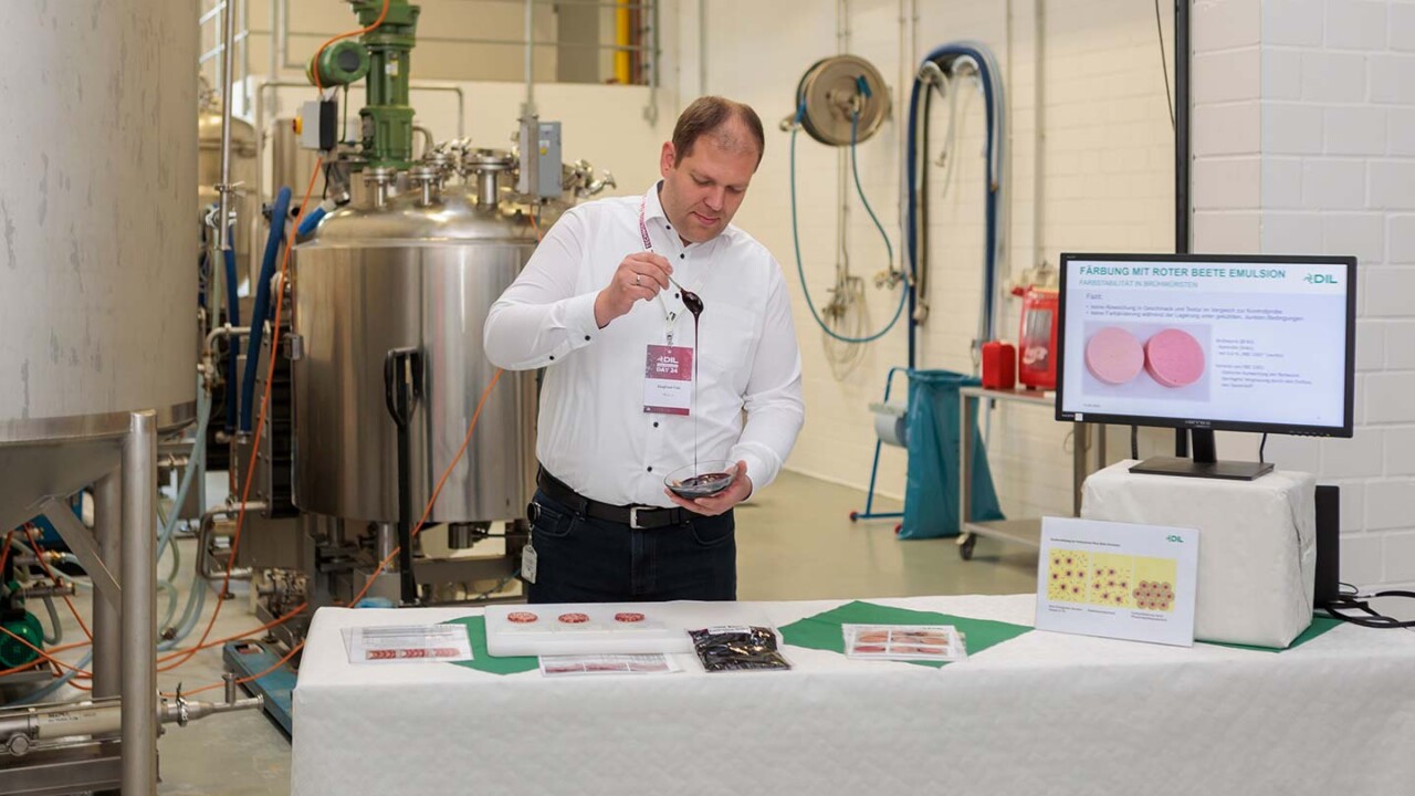 dil-ev-lohnproduktion-disperse-systme-Anwendung-und-Produktdemonstration-am-DIL Ein Mitarbeiter demonstriert in einer Pilotanlage die Verarbeitung bzw. Applikation eines Produkts an einem Probentisch. Im Hintergrund sind Edelstahltanks und Anlagentechnik zu sehen; rechts zeigt ein Monitor eine Präsentationsfolie zur „Färbung mit roter Bete Emulsion“ mit Produktabbildungen. Auf dem Tisch liegen Proben und Informationsmaterial.
