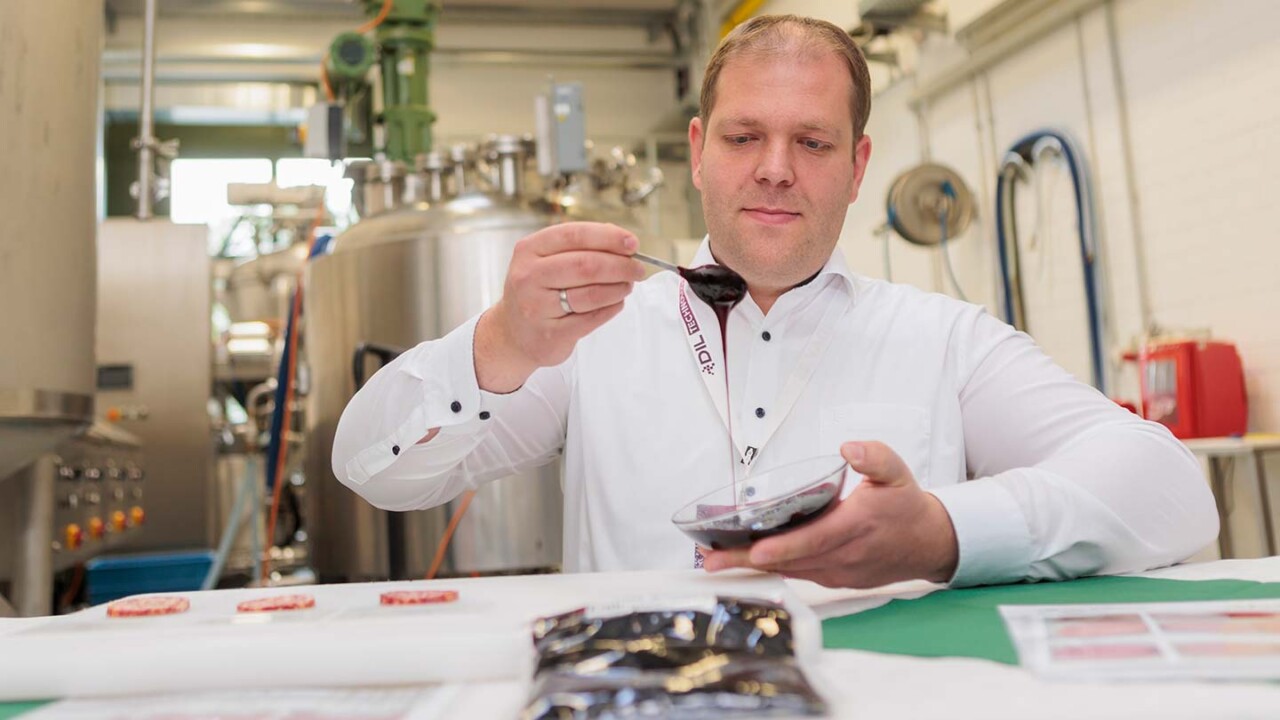 dil-ev-lohnproduktion-disperse-systme-Produktapplikation-im-Technikumsmaßstab-am-DIL Ein Mitarbeiter trägt in einer Pilotanlage eine dunkelrote Emulsion mit einem Löffel auf Lebensmittelproben auf. Im Vordergrund liegen vorbereitete Muster auf einem Tisch, im Hintergrund sind Edelstahltanks und Anlagentechnik einer verfahrenstechnischen Umgebung zu sehen.