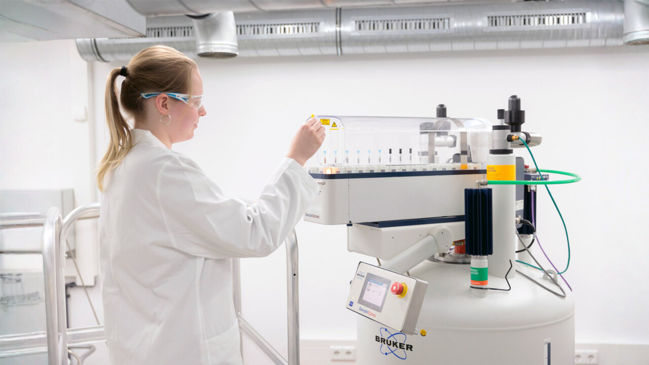 dil-ev-sicherheit-authentizitaet-chemische-analytik-Analytik-Labor Laboraufnahme: Eine Mitarbeiterin in Laborkittel und Schutzbrille bedient einen automatisierten Probenwechsler an einem modernen NMR-System. Das Gerät mit zahlreichen Probenröhrchen steht für präzise chemische Analytik, strukturaufklärende Messverfahren und Qualitätssicherung – z. B. zur Prüfung von Lebensmittelsicherheit und Authentizität.