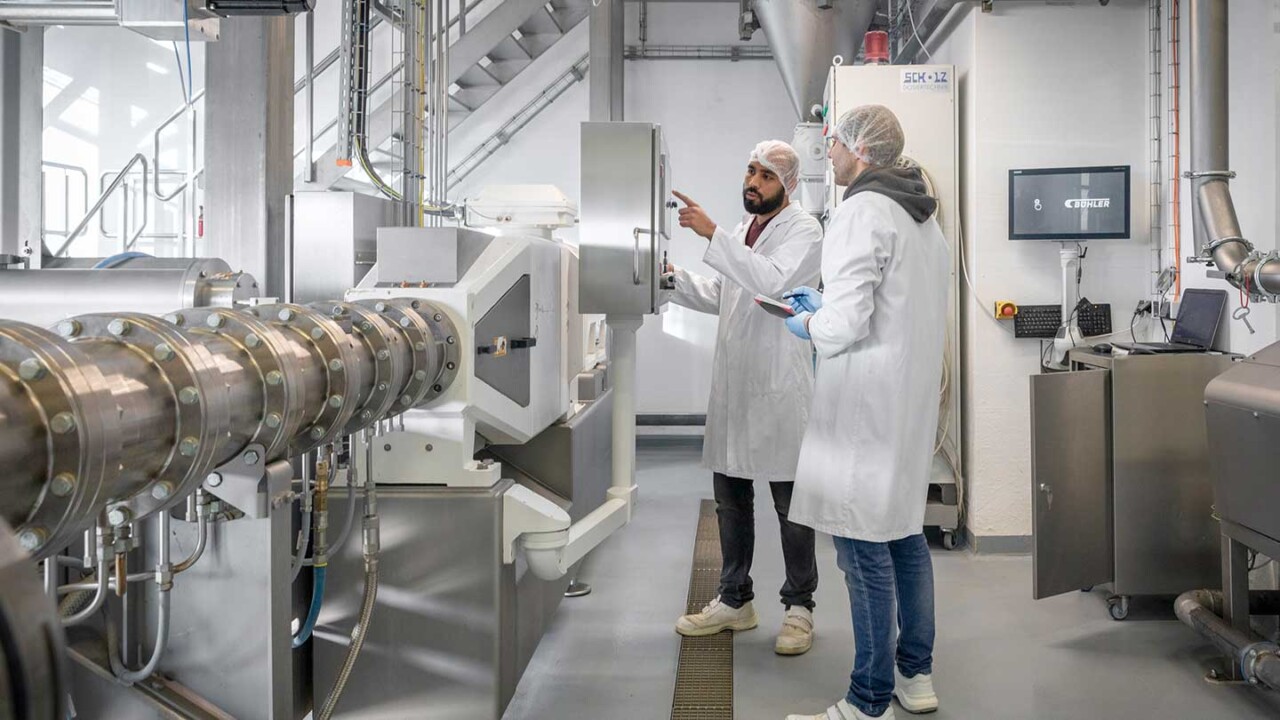 Zwei Mitarbeitende in Hygienekleidung stehen in einer Technikumsanlage und bedienen bzw. überprüfen eine industrielle Prozessmaschine mit Rohrleitungen, Aggregaten und Steuerungseinheit. Einer erklärt Einstellungen am Bedienfeld, der andere dokumentiert die Daten. Das Bild steht für Verfahrenstechnik, Pilotierung, Scale-up, Prozessmonitoring und Qualitätssicherung in der Lebensmittelverarbeitung.