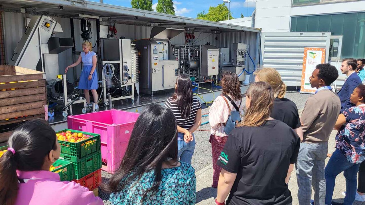 Außenaufnahme bei einer Vorführung am DIL: Eine Gruppe von Teilnehmenden verfolgt eine Präsentation an einer mobilen Container-Prozessanlage. Im Vordergrund stehen Kisten mit Obst, die auf den Praxisbezug zur Lebensmittelverarbeitung hinweisen. Das Bild steht für Technologietransfer, Advanced Technologies, Pilotierung und Demonstration neuer Verfahren in der Lebensmitteltechnik – von Rohware bis Prozessanlage.