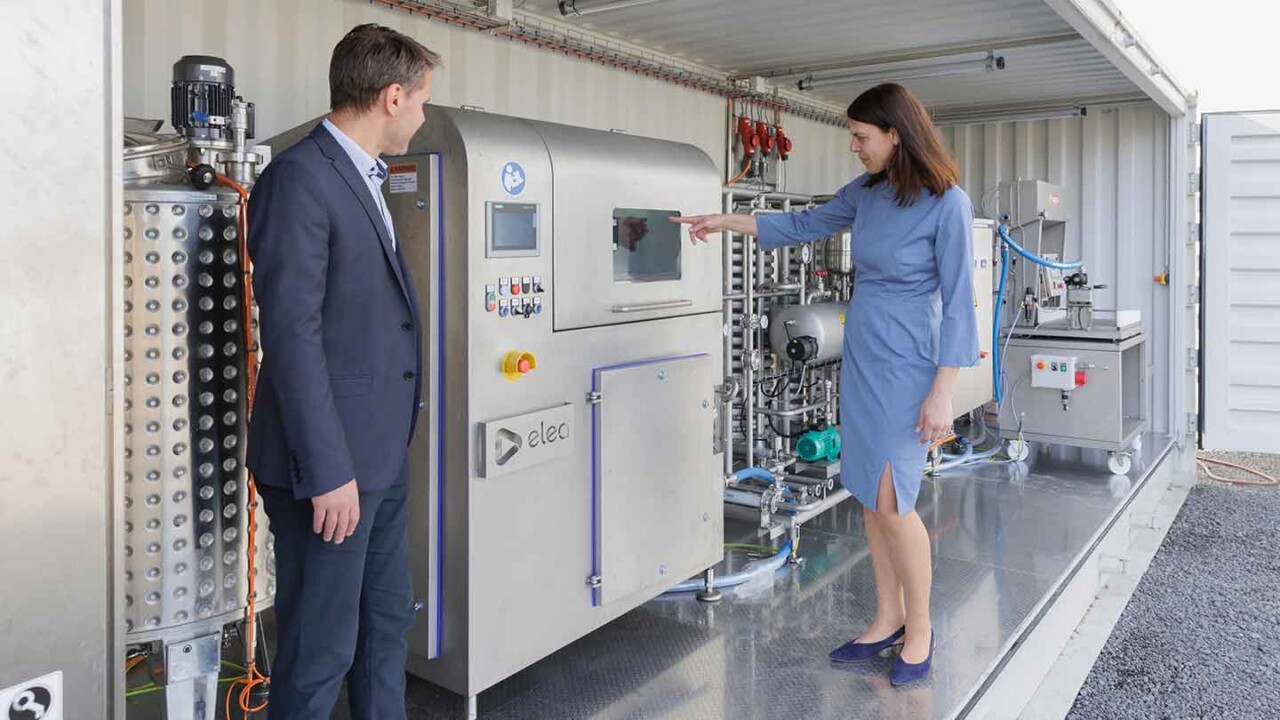 Zwei Personen stehen in einer mobilen Container-Pilotanlage und besprechen eine industrielle Prozessanlage aus Edelstahl. Eine Frau zeigt auf das Bedienfeld der Anlage, während ein Mann im Anzug zuhört. Das Bild steht für Technologietransfer, Prozessentwicklung und Demonstration moderner Verfahrenstechnik im Pilotmaßstab – z. B. für Anwendungen in der Lebensmittelverarbeitung und „Advanced Technologies“.