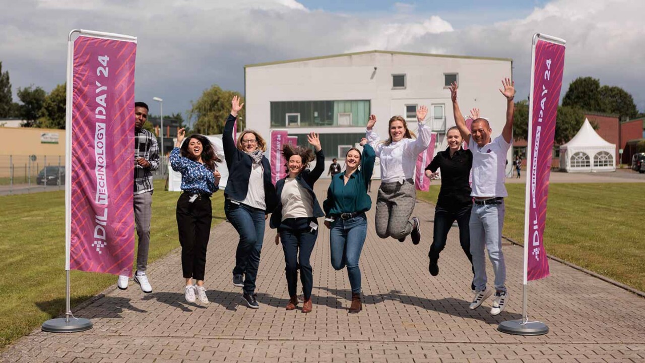 Gruppenfoto im Außenbereich: Mitarbeitende springen und jubeln zwischen zwei Beachflags mit der Aufschrift „DIL Technology Day 24“. Im Hintergrund ist ein Institutsgebäude zu sehen. Das Bild vermittelt Teamgeist und Veranstaltungsatmosphäre und steht für Austausch, Innovation und Vernetzung rund um Forschung und Lebensmitteltechnologie.