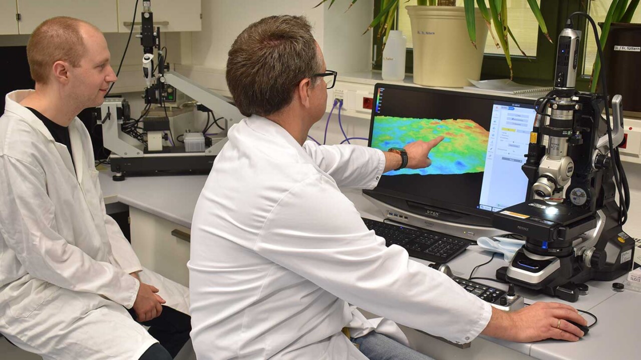 dil-ev-struktur-verarbeitung-struktur-und-funktionalitaet-Mikrostruktur-und-Oberflaechenmessung-mit-Digitalmikroskop Zwei Wissenschaftler im Laborkittel werten am Monitor eine farbcodierte Oberflächen- bzw. Höhenkarte einer Probe aus. Neben dem Bildschirm steht ein Digitalmikroskop zur hochauflösenden Bildaufnahme. Das Foto steht für Strukturanalyse, Mikrostruktur-Charakterisierung, Struktur-Funktionalitätsforschung und die Bewertung von Verarbeitungseinflüssen auf Produkteigenschaften – z. B. in der Lebensmittel- und Materialforschung.