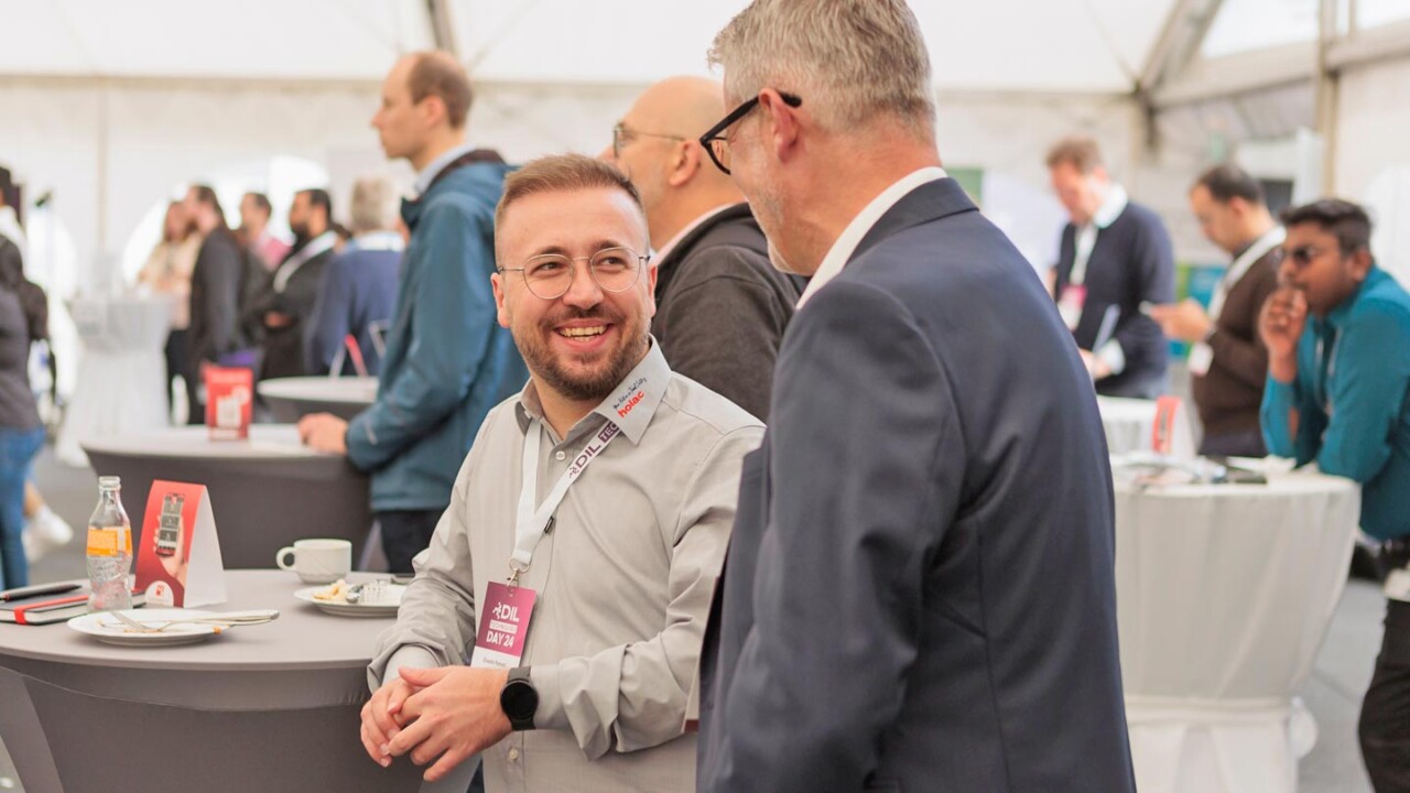 In einem Veranstaltungszelt unterhalten sich zwei Teilnehmer an einem Stehtisch. Im Vordergrund lächelt ein Mann mit Brille und Event-Badge („DIL DAY 24“) im Gespräch mit einem weiteren Teilnehmer im Anzug. Im Hintergrund sind weitere Gäste, Stehtische und Catering-Situation zu sehen – typische Networking-Atmosphäre bei einer Fachveranstaltung.