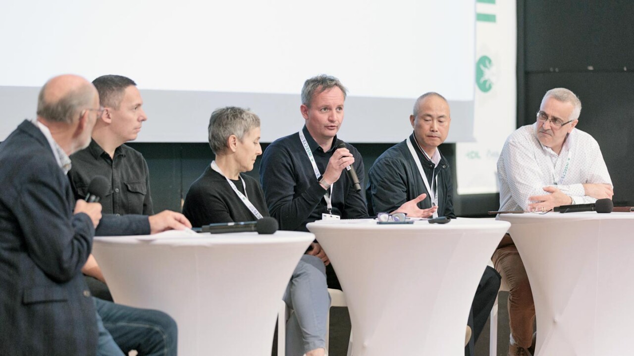 Mehrere Fachleute sitzen auf einer Bühne an Stehtischen und diskutieren im Rahmen einer Podiumsrunde. Ein Sprecher hält ein Mikrofon und erläutert einen Punkt, während die übrigen Panelteilnehmenden aufmerksam zuhören. Die Szene zeigt eine professionelle Konferenzsituation mit Fokus auf fachlichen Austausch, aktuelle Entwicklungen und Perspektiven rund um Insekten als nachhaltige Proteinquelle und Innovationen für die Lebensmittel- und Feed-Branche.